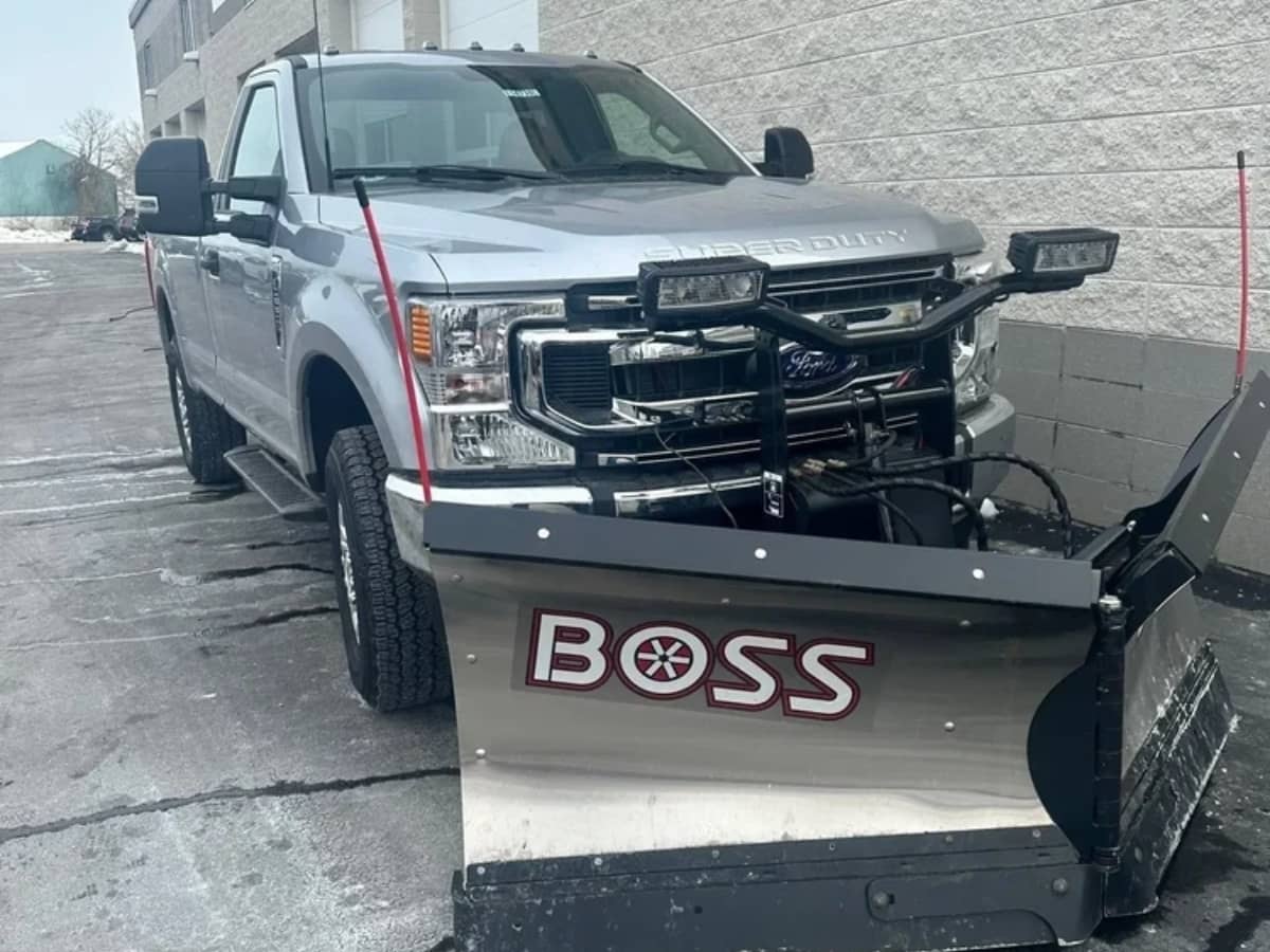 Silver Ford Super Duty pickup truck with a large snow plow attached to the front, parked outside on a slightly snowy and icy pavement next to a gray brick building.